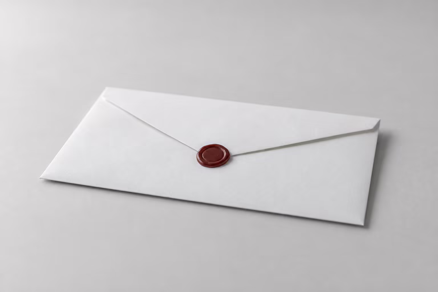 A plain white envelope lying sealed on a grey surface, one small dark red wax dot at the seal